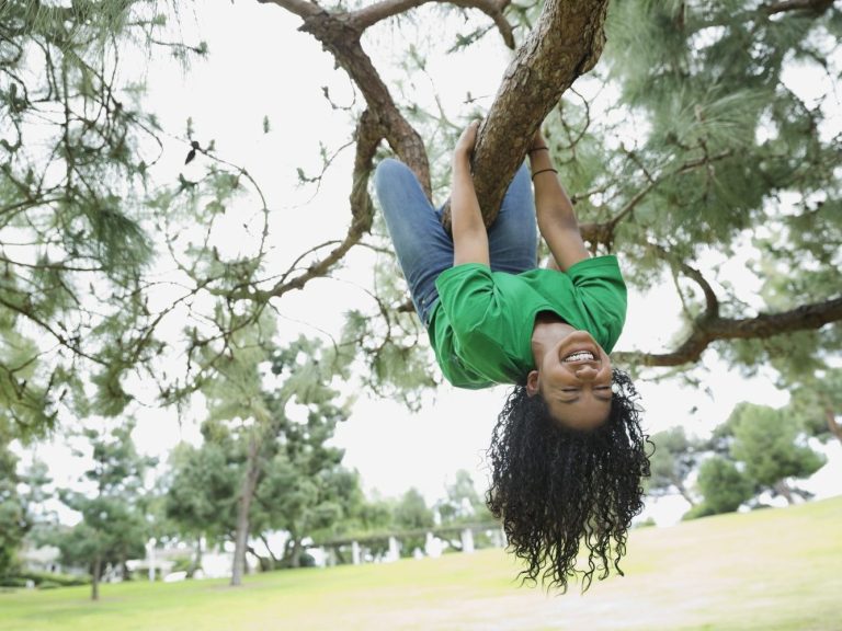 A girl hanging upside down from a tree branch, smiling amidst greenery.