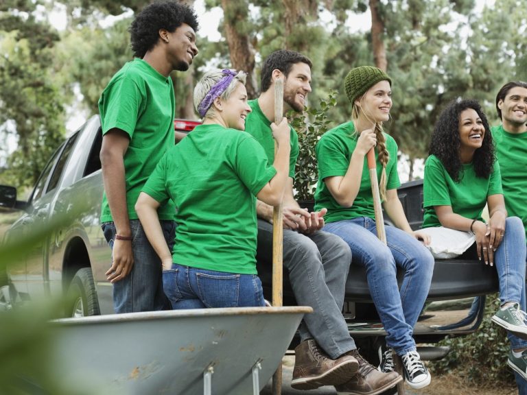 A diverse group of six smiling volunteers in green shirts, sitting on a pickup truck.